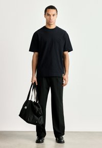 Μαύρο μπλουζάκι με λαιμόκοψη crew και τσέπη, συνδυασμένο με χαλαρούς μαύρους παντελόνες και μαύρη τσάντα duffle με λευκό κείμενο και πολύχρωμες ρίγες.