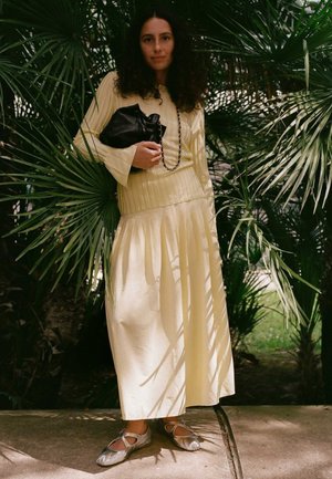 Kvinde med krøllet mørkt hår iført en lysegul ribbet top og lang nederdel, holder en sort håndtaske, stående blandt grønne palmeblade.