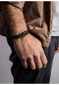 Braided brown leather bracelet with a black metal accents, worn on a wrist, paired with a brown corduroy shirt and dark pants.