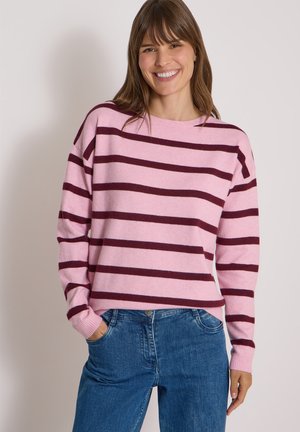 Femme souriante portant un pull rose avec des rayures horizontales foncées et un jean bleu, la main gauche dans sa poche.