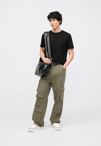 T-shirt preta de algodão combinada com calças cargo verde-oliva com múltiplos bolsos. Modelo carrega uma bolsa preta ao ombro e usa sapatilhas brancas.