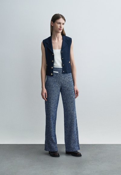 Γυναίκα όρθια, φορούσα κοντό γιλέκο με κουμπιά σε ναυτικό μπλε, λευκό τοπ, μπλε υφασμάτινη παντελόνα με φαρδιά πόδια και μαύρα παπούτσια, με φόντο απλό ανοιχτόχρωμο.