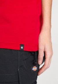 T-shirt en coton rouge avec ourlet cousu et étiquette de logo DC en bas. Assorti à un pantalon sombre arborant un logo de marque visible.