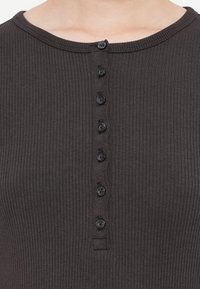 Close-up van de ribbelige, donkergrijze voorkant van een shirt met een ronde halslijn en zeven zwarte knopen die verticaal in het midden zijn uitgelijnd.