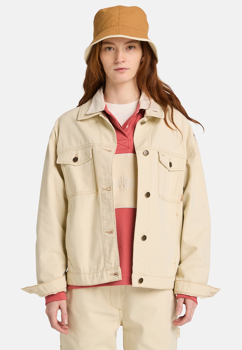 Veste en denim beige avec poches avant, col beige et boutons en cuivre, portée sur un polo rose et crème assorti à un pantalon, accompagnée d'un bob marron.