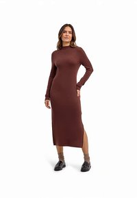 Langes, schokoladenbraunes Strickkleid mit hohem Kragen, langen Ärmeln, Seitenschlitzen und einer figurbetonten Silhouette. Kombiniert mit schwarzen Schuhen und hellen Socken.