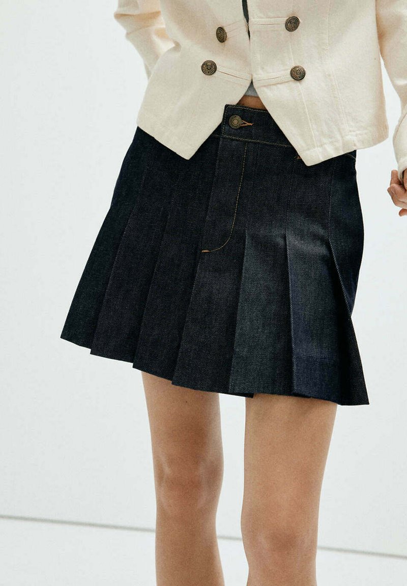 Gros plan d'une personne portant une jupe plissée sombre et une veste de couleur crème avec des boutons décoratifs sur un fond clair uni.