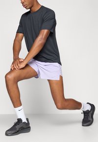 Schwarzes kurzärmeliges Shirt, helllila Shorts, schwarze Sneaker und weiße Socken. Das Subjekt befindet sich in einer Ausfallschritt-Position und zeigt Details von Beinen und Armen.