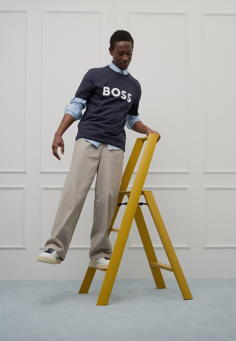 T-shirt navy à manches courtes avec le texte "BOSS" en blanc, pantalon beige, debout sur une échelle pliante jaune vif contre un fond clair.