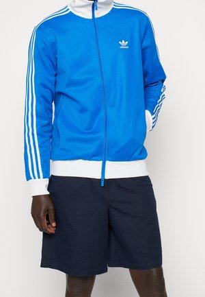 Osoba ubrana w niebieską kurtkę Adidas na zamek z białymi paskami na rękawach i ciemnogranatowe spodenki, stojąca na jednolitym tle.