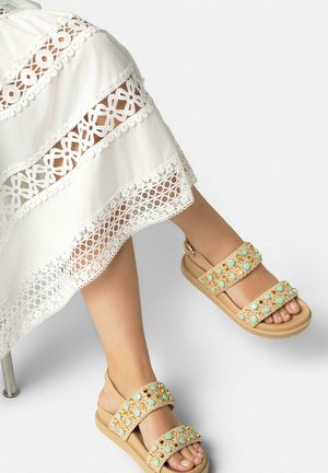Füße in beigen Sandalen mit türkisfarbenen Steinen und goldenen Verzierungen, kombiniert mit einem weißen Spitzenkleid mit floralen und kreisförmigen Mustern.