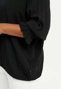 Blusa nera con un accento arricciato al polsino, realizzata in morbido tessuto con una vestibilità rilassata. Etichetta visibile all'orlo.