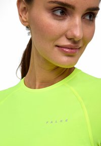 Woman with tied-back hair wearing a bright neon green shirt with "FALKE" logo, looking to her side with a slight smile.