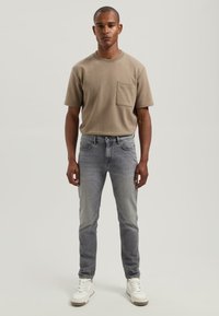 Beige színű pamut póló, mellzsebkészítménnyel, világosszürke, slim fit farmernadrággal párosítva, valamint fehér sneakers cipőkkel, amelyek textúrázott talppal rendelkeznek.