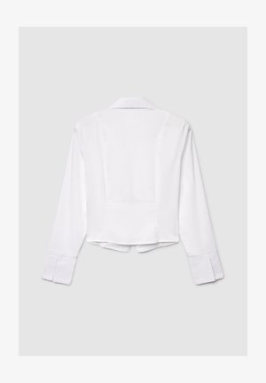 Chemisier blanc court avec manches longues, col et poignets boutonnés. Tissu lisse avec un design épuré et simple, et détail de couture au dos.