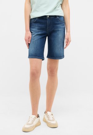Pantalones cortos de mezclilla en azul oscuro con corte recto, que cuentan con cinco bolsillos y dobladillos enrollados. Llevados con zapatillas de color claro.