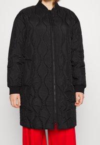 Veste longue matelassée noire avec une fermeture éclair à l'avant, des poignets côtelés et des poches latérales. Présente un motif texturé et une coupe décontractée.