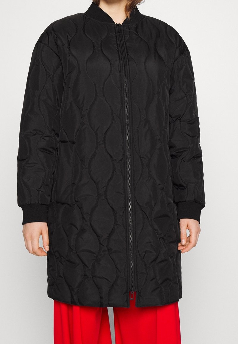 Veste longue matelassée noire avec une fermeture éclair à l'avant, des poignets côtelés et des poches latérales. Présente un motif texturé et une coupe décontractée.
