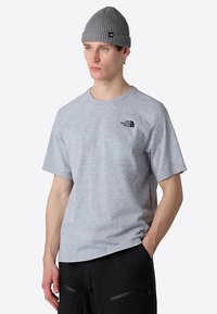 Szary bawełniany T-shirt z krótkimi rękawami i okrągłym dekoltem, z małym czarnym logo na piersi. Noszony z czarnymi spodniami i szarą czapką beanie.