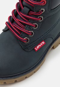 Tmavě zelená bota s červenými a modrými pruhovanými tkaničkami, kovovými dírkami na tkaničky a červenou štítkem s logem Levi's. Texturovaný svršek a robustní béžová podrážka.