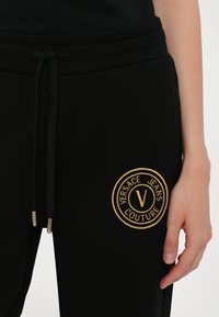 Črne jogger hlače iz mehkega blaga s pasom na vrvico. Imajo krožno, zlatoprevedeno logo "Versace Jeans Couture" na stegnu.