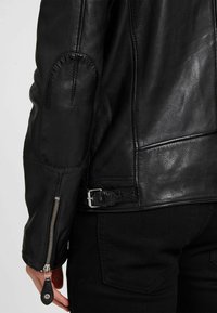 Schwarze Lederjacke mit Reißverschlussärmeln, geschwungenen Schulternähten und Schnallen-Detail an der Taille. Glatte Textur und taillierte Passform.