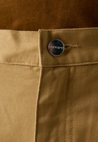 Khakihosen aus einem strukturierten Stoff, mit einem Metallknopf mit "Carhartt"-Gravur und sichtbaren Nähten.