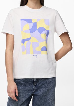 Persoon draagt een wit T-shirt met korte mouwen met abstracte gele en paarse vormen en een blauwe spijkerbroek, staand met één arm achter de rug gebogen.