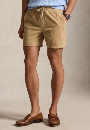 Homme portant un short beige à cordon, un pull en maille bleu clair, des mocassins en cuir marron et une montre, debout devant un fond neutre.