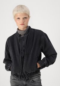 Mörkgrå denimjacka med croppad design, tryckknappsstängning, elastiska ärmslut och två fickor framtill. Bärs över en grå turtleneck.