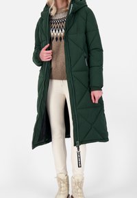 Lång grön vadderad kappa med kviltad design, huva, sidofickor och hel dragkedja. Bärs med krämfärgade byxor och mönstrad tröja.