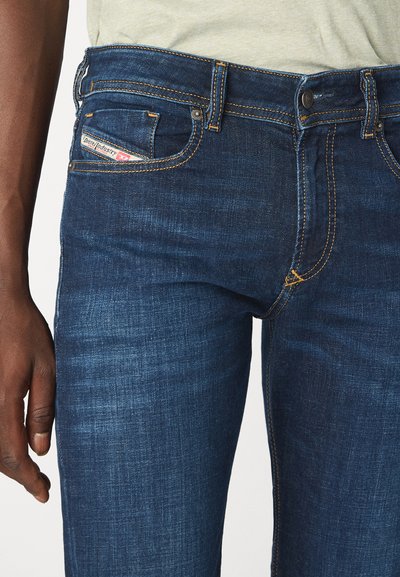 Jeans en denim bleu foncé avec des coutures jaunes contrastées, design à cinq poches et un petit logo sur la poche avant. Texture douce.