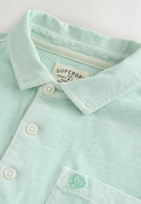 Polo vert clair avec col boutonné, trois boutons blancs, poche poitrine et étiquette "Superdry Co" sur le col intérieur.