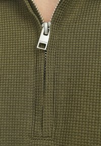 Olivgrön zip-up tröja med en texturerad, våffelstickad tyg. Har en silverfärgad dragkedja och en märkesdragflik för extra detalj.