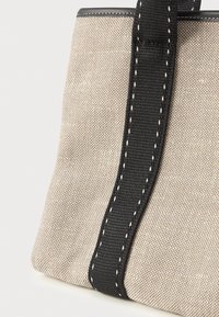 Μπεζ καμβάς τσάντα tote με υφή, διαθέτοντας μαύρη λαβή με λευκές ραφές και αντίθετο άκρο στο πάνω μέρος.