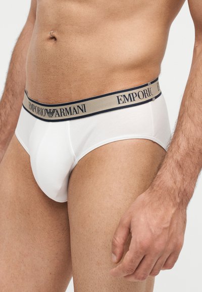 Αρσενικός κορμός που φοράει λευκά εσώρουχα Emporio Armani με μπεζ και ναυτικό μπλε λάστιχο στη μέση, δείχνοντας μέση, γοφούς και δεξί χέρι.