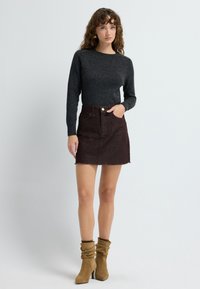 Vrouw met krullend haar in een donkergrijze trui, een bruine spijkerrok met een rauwe zoom en beige slouchy haklaarzen, staande tegen een witte achtergrond.