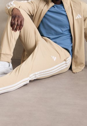 Osoba ubrana w beżowy dres Adidas z białymi paskami i niebieską koszulę, siedząca na jasnoszarej podłodze przy neutralnej ścianie.
