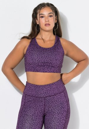 Jonge vrouw met lang gevlochten haar, die oorringen draagt en een bijpassende paarse sportbeha en legging met luipaardprint, staand met de handen in de zij.