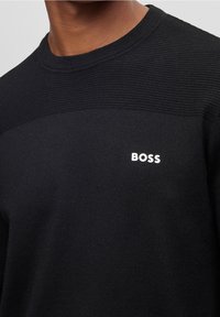 Svart tröja med en texturerad överdel, ribbad halsringning och en vit "BOSS"-logotyp på bröstet. Tillverkad av ett mjukt stickat material.