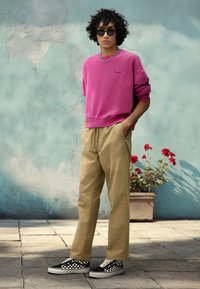 Rosa sweatshirt med broderet logo, beige bukser og sort-hvide ternede sneakers. Modellen bærer solbriller og en kæde halskæde.