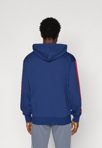 Ναυτικό μπλε φούτερ με υφή, που διαθέτει μεγάλη κουκούλα και κόκκινες πλευρικές ρίγες. Ο σχεδιασμός περιλαμβάνει ριμπ μανσέτες και στρίφωμα για καλύτερη εφαρμογή.