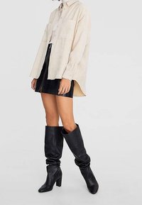 Camicia oversize beige chiaro con bottoni, minigonna in velluto nero, e stivali neri in pelle con tacco al ginocchio, con un design morbido.