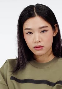 Portrét osoby s dlhými čiernymi vlasmi, ktorá nosí zelenú mikinu s čiernym horizontálnym prúžkom cez hruď.