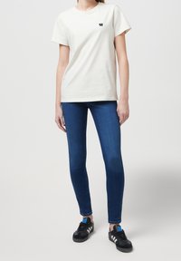 Camiseta blanca de manga corta con un pequeño logo oscuro en el pecho, combinada con jeans skinny azules y zapatillas negras con acentos azules.