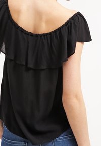 Top negro sin mangas con detalle de volantes, hecho de tejido ligero. Presenta un ajuste relajado y un escote fruncido. Llevado con jeans de mezclilla azul.