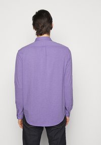 Košile s dlouhým rukávem, s knoflíky, v jasně fialovém materiálu. Obsahuje klasický límeček, viditelné švy a zakřivený lem.