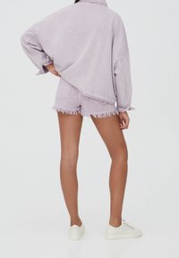 Mujer de pie con una mano en la cadera, vistiendo una chaqueta de mezclilla oversized de color púrpura claro y shorts deshilachados, con zapatillas blancas, sobre un fondo blanco.