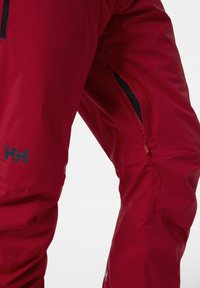 Červené vodotěsné outdoorové kalhoty s kapsou na zip, hladkým povrchem a logem "HH" na dolní části nohy. Navrženy pro funkčnost.