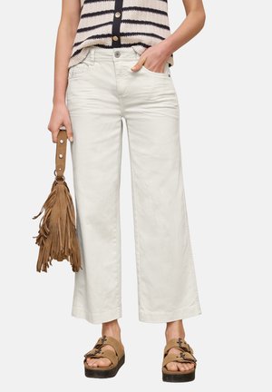 Kvinde iført hvide bukser med vidde ben, striped ærmeløs top, tan frynse håndtaske og tan spænde sandaler, står med den ene hånd i lommen.